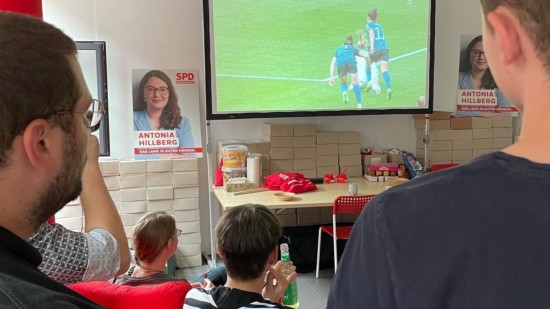 Eine Leinwand in einem Raum, auf der Fußball läuft. Im Raum stehen und sitzen Menschen die das Spiel schauen.