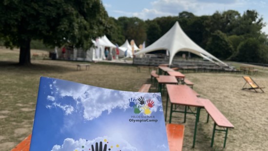 Programmheft des Olympiacamps, im Hintergrund Zelte und Sitzgelegenheiten.